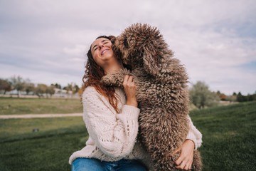 A beautiful woman is in the meadow with her dog. The owner is hugging her pet while the dog is licking her face.They are enjoying a day in the park. The pet is a Spanish water dog with brown fur.
