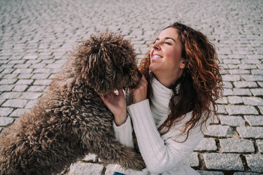 A beautiful woman is in the park with her dog. The owner is playing with her pet while the dog is licking her hand.They are enjoying a day together. The pet is a Spanish water dog with brown fur.