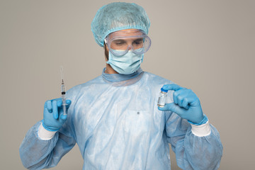 Doctor in medical mask and protective googles holding jar of vaccine and syringe isolated on grey
