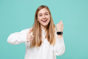 Cheerful young woman in casual white hoodie posing isolated on blue turquoise background studio portrait. People lifestyle concept. Mock up copy space. Wearing smart watch on hand, showing thumb up.