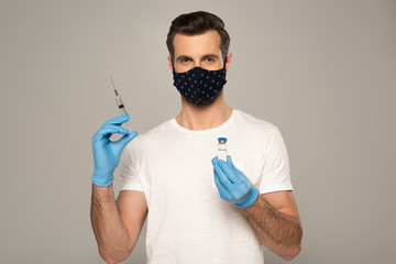 Man in safety mask and latex gloves holding jar of vaccine and syringe isolated on grey