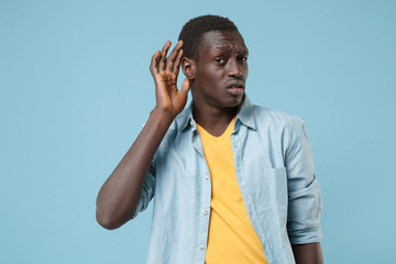 Bewildered young african american man guy in casual shirt, yellow t-shirt posing isolated on blue background. People lifestyle concept. Mock up copy space. Try to hear you with hand gesture near ear.