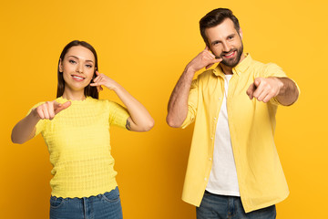 Positive couple pointing with fingers at camera and showing call gesture on yellow background