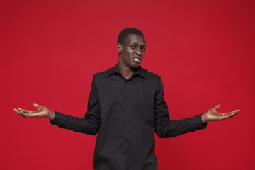 Perplexed puzzled young african american man in classic black shirt posing isolated on red background studio portrait. People sincere emotions lifestyle concept. Mock up copy space. Spreading hands.