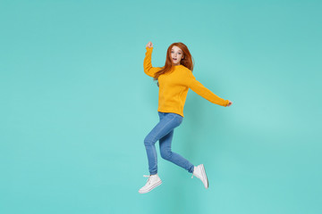 Fototapeta na wymiar Shocked young redhead woman girl in yellow knitted sweater posing isolated on blue turquoise background studio portrait. People emotions lifestyle concept. Mock up copy space. Jumping spreading hands.