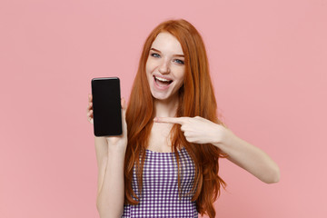 Excited young redhead woman girl in plaid dress posing isolated on pastel pink background. People lifestyle concept. Mock up copy space. Pointing index finger on mobile phone with blank empty screen.