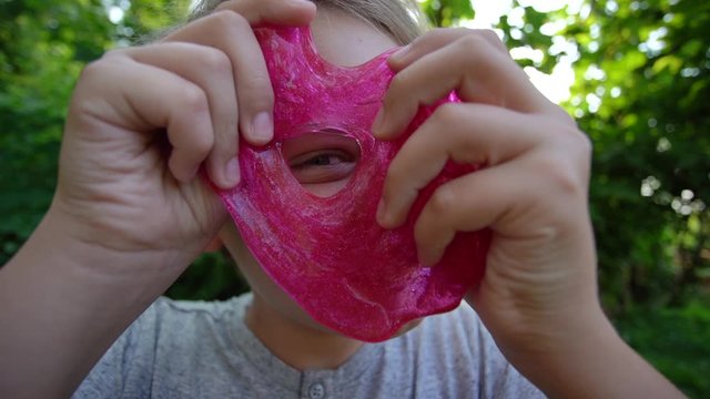 Closeup view video portrait of cute funny face of white kid playing cheerfully outdoor and looking through circle made of pink transparent slime toy.