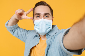 Close up of man in sterile face mask isolated on yellow background. Epidemic pandemic coronavirus 2019-ncov sars covid-19 flu virus concept. Doing selfie shot on mobile phone showing victory sign.
