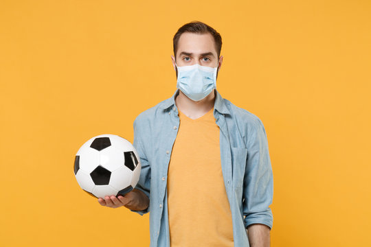 Young man in face mask isolated on yellow background. Epidemic pandemic 2019-ncov sars covid-19 flu virus concept. Coronavirus stops football sport. Cheer up support favorite team with soccer ball.