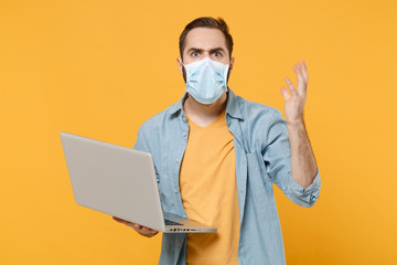 Irritated young man in sterile face mask posing isolated on yellow background. Epidemic pandemic coronavirus 2019-ncov sars covid-19 flu virus concept. Working on laptop pc computer spreading hands.