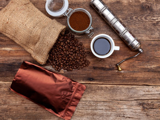 Fototapeta na wymiar Bag of coffee on a wooden table. Coffee beans ground in a jar. Manual coffee grinder. Top view, place for text. Brown background. Packing coffee in a pack. White up with a drink.