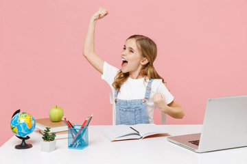 Happy little kid schoolgirl 12-13 years old study at white desk with pc laptop isolated on pink background. School distance education at home during quarantine concept. Clenching fists like winner.