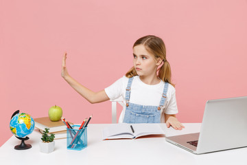 Serious little kid schoolgirl 12-13 years old study at desk with laptop isolated on pink background. School distance education at home during quarantine concept. Showing stop gesture with palm aside.