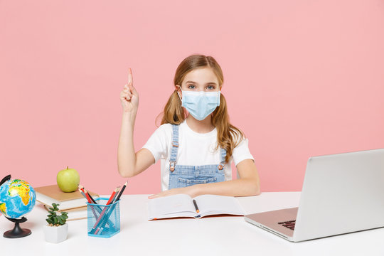 Little Kid Schoolgirl 12-13 Years Old In Face Mask Study At Desk With Pc Laptop Isolated On Pastel Pink Background. School Distance Education At Home During Quarantine Concept. Point Index Finger Up.