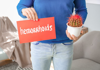 Young man holding paper with text HEMORRHOIDS and cactus at home