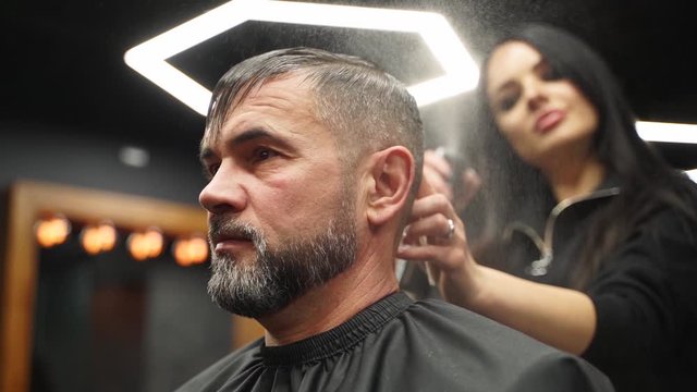 Barber sprays water on client hair before haircut and styling in barbershop. Hairdresser woman wets hair by spray and combs them in hair salon. Master use water sprayer bottle in male beauty studio.