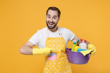 Excited young man househusband in apron rubber gloves doing housework isolated on yellow...
