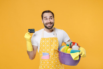 Pensive young man househusband in apron rubber gloves hold basin with detergent bottles washing...