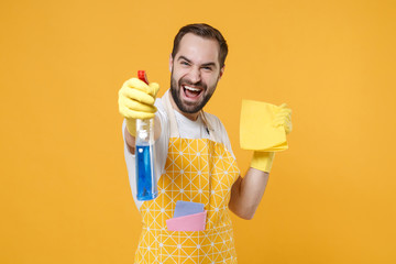 Laughing young man househusband in apron rubber gloves hold spray with washing cleanser, cleaning...