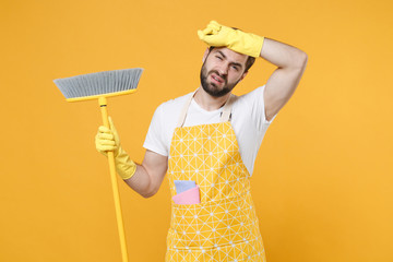 Exhausted young man househusband in apron rubber gloves hold in hands broom while doing housework isolated on yellow wall background studio portrait. Housekeeping concept. Put hand on head.