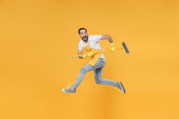 Fototapeta na wymiar Side view of excited crazy young man househusband in apron rubber gloves hold in hands broom while doing housework isolated on yellow background studio. Housekeeping concept. Jumping, looking camera.