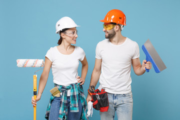 Smiling couple woman man in protective helmet hardhat hold paint roller for painting, putty knife isolated on blue background. Instruments accessories renovation apartment room. Repair home concept.