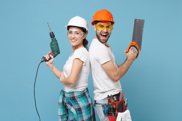 Excited couple woman man in protective helmet hardhat hold electric drill saw isolated on blue...