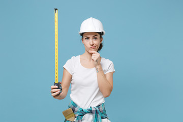 Confused young woman in protective helmet hardhat hold measure tape isolated on blue background. Instruments accessories for renovation apartment room. Repair home concept. Put hand prop up on chin.