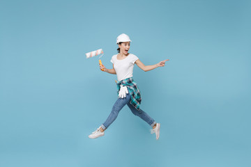Plakat Side view excited woman in helmet hardhat jump with paint roller isolated on blue background. Instruments accessories for renovation apartment room. Repair home concept. Pointing index finger aside.