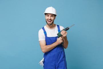 Laughing young man in coveralls protective helmet hardhat hold electric drill isolated on pastel...