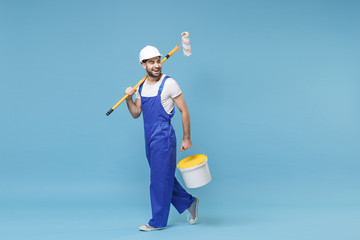 Cheerful young man in coveralls protective helmet hardhat hold paint roller, bucket isolated on pastel blue wall background. Instruments accessories for renovation apartment room. Repair home concept.