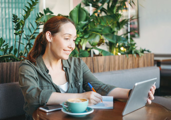 Adult smiling brunette woman doing notes and look at tablet takes online course on table at cafe