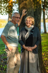 An elderly man with a mustache and glasses stands with an elderly woman grandmother on a summer evening in the city Park by the river and smile