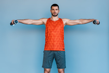 Naklejka premium bearded sportsman standing with outstretched hands and exercising with dumbbells on blue