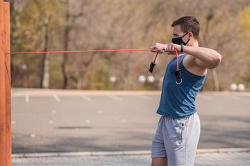 A young and athletic guy in a medical mask does exercises with sports elastic during a pandemic....