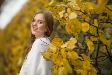 Beautiful happy girl with light brown hair developing in the wind among yellow foliage in an autumn park. Attractive young woman in a white sweater smiles joyfully. Fall season. Amazing autumn mood.