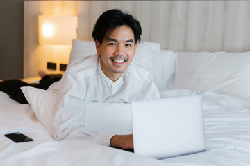 Asian male in white shirt using laptop while working on bed. work from home concept.