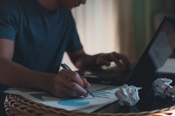 A man hand holding pen with graph and business documents on office table and working on laptop computer.