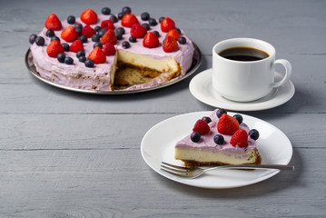 Piece of home made Cheesecake with berries on plate and mug of Americano coffee. Cheesecake with Strawberry, blueberry and raspberry on grey wooden background.