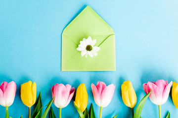 top view of blooming yellow and pink tulips near green envelope with chrysanthemum flower on blue, mothers day concept