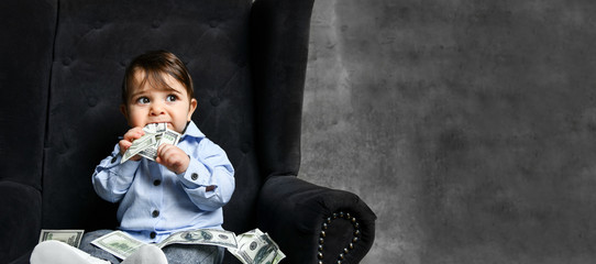 Toddler in blue shirt, gray pants, booties. He holding money, chewing it, sitting in black armchair on gray background. Close up