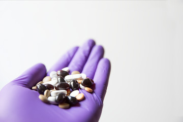 Close up of medical 
violet latex glove with several tablets of different color, shape e dimension. White uniform paper background. Idea of addiction to drugs.