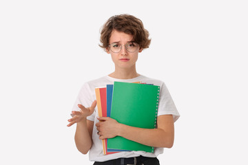 Unhappy young student in a white shirt and eyeglasses holding colorful folders showing so-so gesture