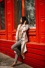 
Street fashion portrait of a young elegant luxurious lady in a beige trench coat, fashion sunglasses, shawl, posing on the street of a European city, against the background of a red building