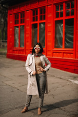 
Street fashion portrait of a young elegant luxurious lady in a beige trench coat, fashion sunglasses, shawl, posing on the street of a European city, against the background of a red building