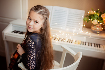 Cute girl with a violin in her hands sits near the piano, bokeh from a garland. Music concept