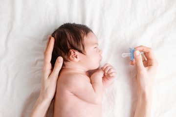 Baby with pacifier on white bed, woman holding nipple in hand