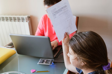 mother working remotely at home with laptop and office document folders while attending to her daughter showing her homework sitting together by covid-19 coronavirus confinement