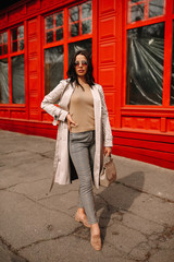 
Street fashion portrait of a young elegant luxurious lady in a beige trench coat, fashion sunglasses, shawl, posing on the street of a European city, against the background of a red building