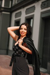 
Beautiful brunette in black overalls, suit, posing on a city street, against the background of a new modern building in dark colors